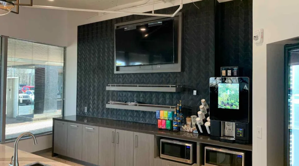 Modern office kitchenette with TV, coffee machine, microwave ovens, and sleek dark cabinetry under natural light.