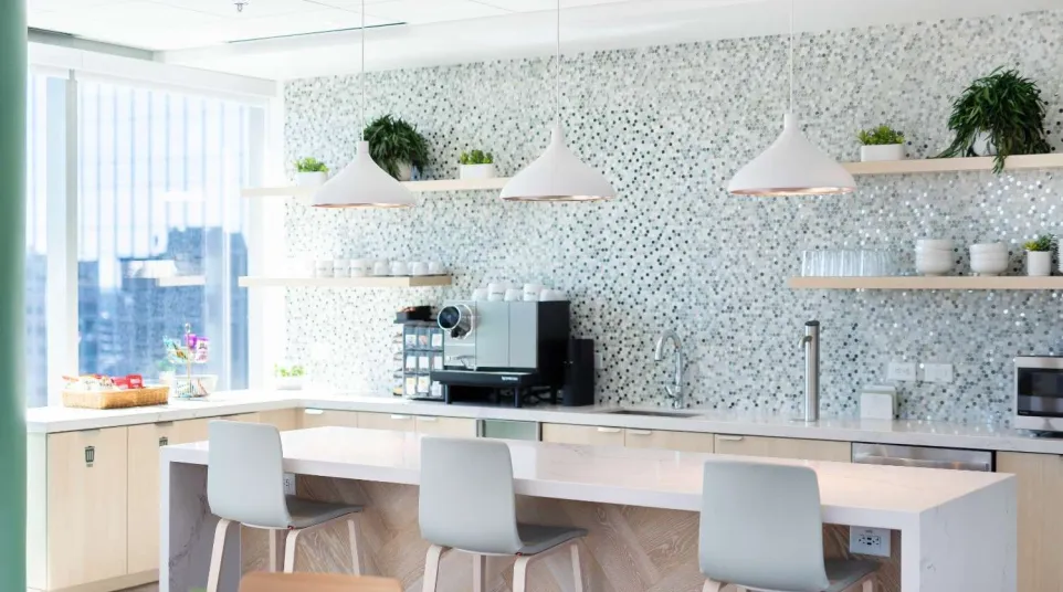 Modern office kitchen with bar stools, white pendant lights, coffee machine, and mosaic tile backsplash.