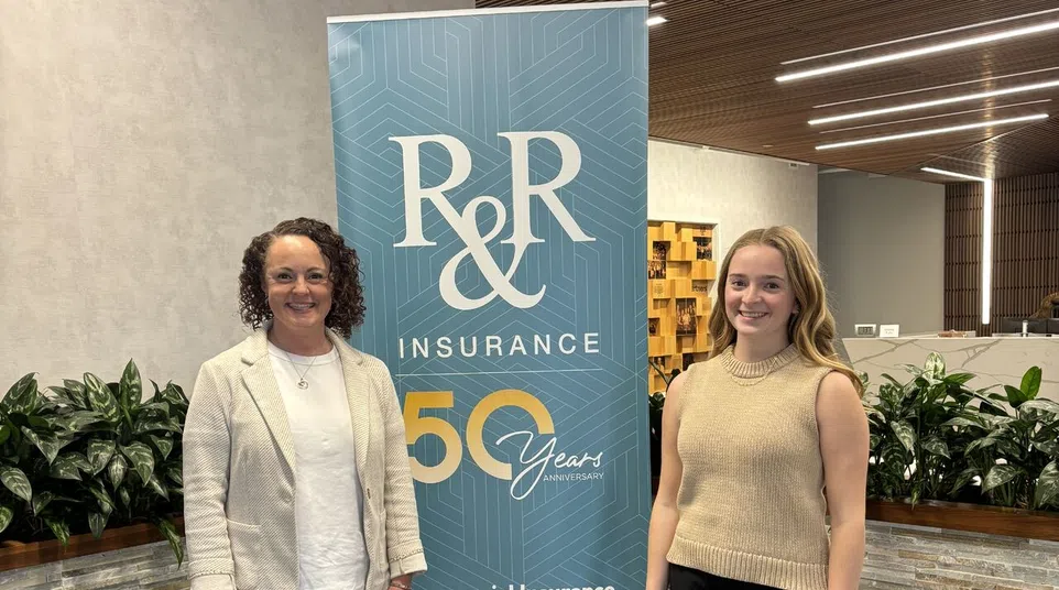 Two women standing beside an R&R Insurance 50 years anniversary banner in a modern office lobby.
