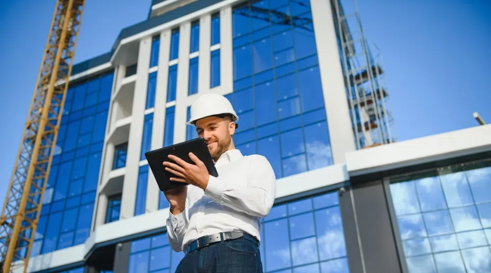 Smiling male architect wearing white hard hat using tablet at modern construction site with glass building.