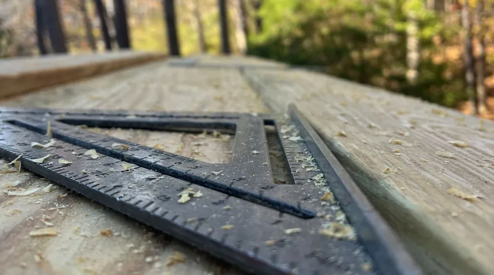 Close-up of a black carpenter's square on a wooden plank with sawdust in an outdoor wooded area.