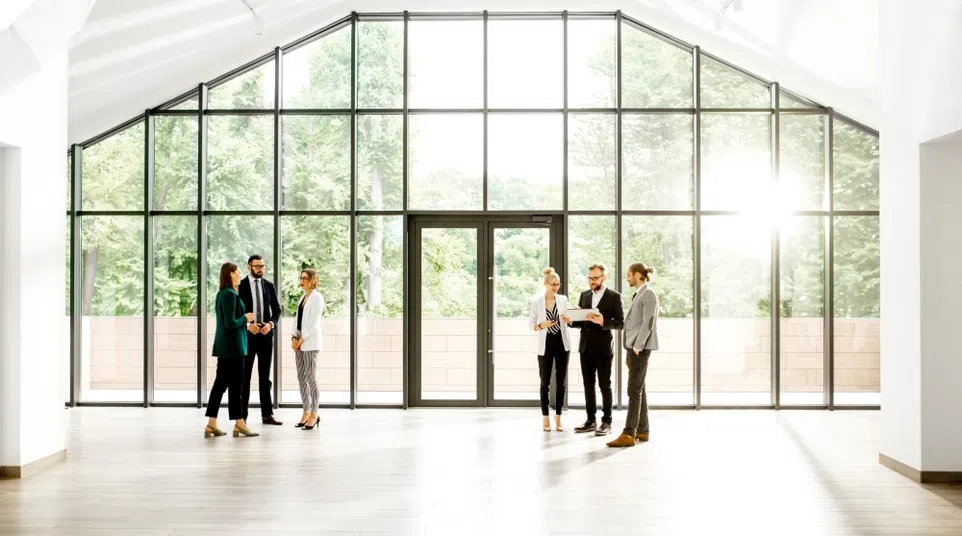 Six business professionals standing and talking in a bright modern office with large floor-to-ceiling windows and natural light.