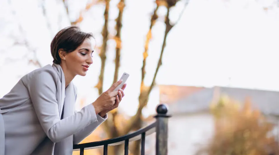Woman in a gray coat leaning on a black iron fence using a smartphone outdoors during autumn.