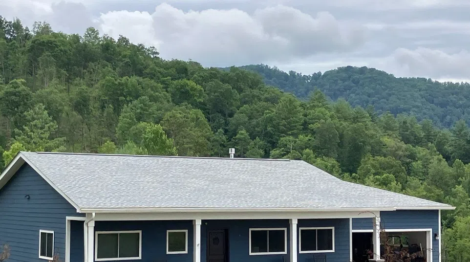 Blue single-story house with white trim and front porch surrounded by green trees and hills under cloudy sky