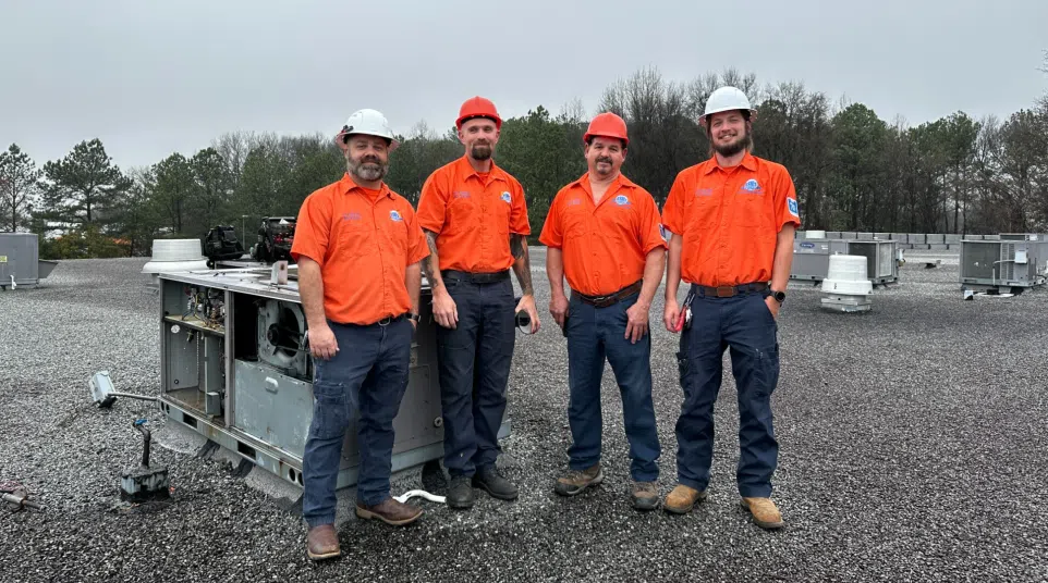Four HVAC technicians in orange uniforms and hard hats standing on a rooftop beside HVAC equipment on a cloudy day
