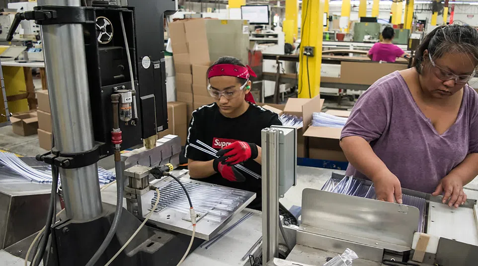 Factory workers assembling metal parts on a production line with machinery and packing boxes in the background