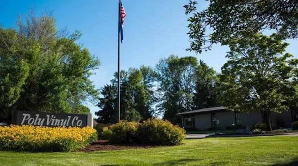 American flag flying on flagpole near Poly Vinyl Co sign surrounded by bushes and green trees on sunny day