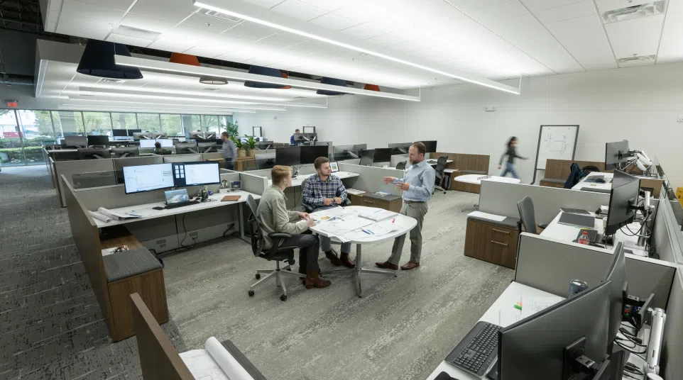 Modern open office space with employees collaborating at a round table surrounded by workstations and computers.
