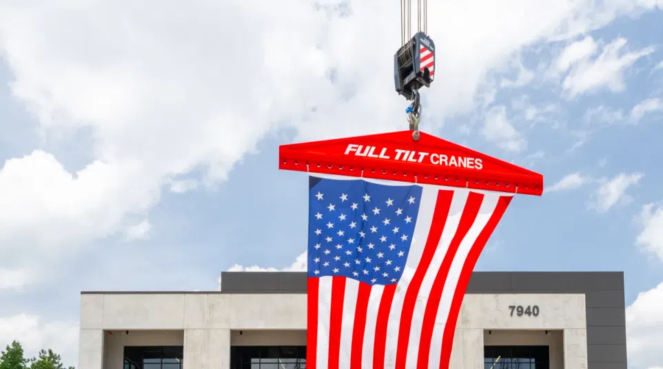 American flag hanging from a Full Tilt Cranes hook above trucks in front of a modern industrial building.