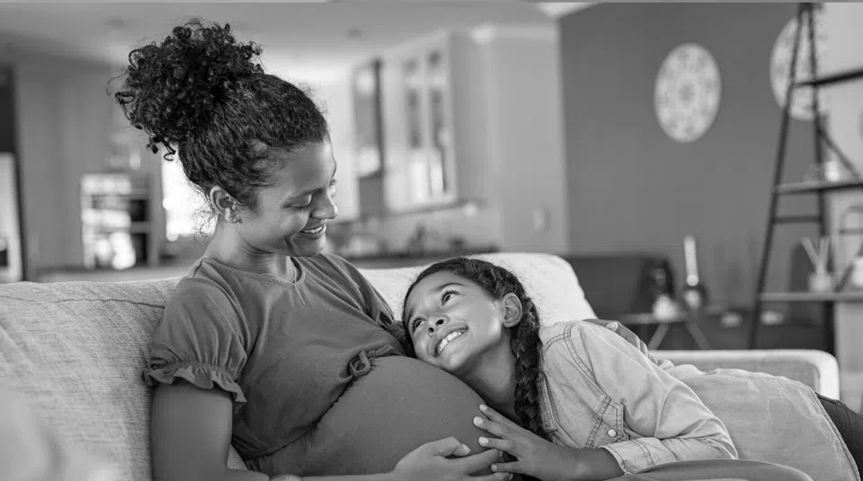 Smiling pregnant woman sitting on couch with daughter resting head on her belly in cozy home setting