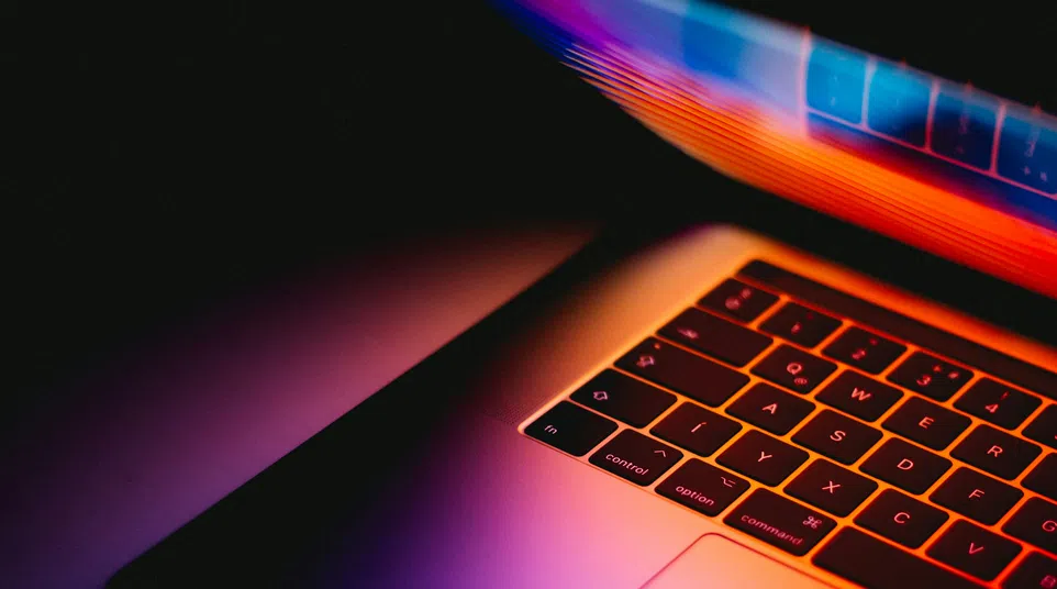 Partial view of a laptop keyboard illuminated by colorful neon lights in a dark environment.