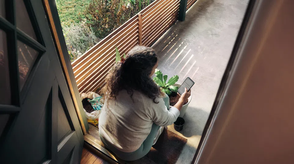 Woman sitting on doorstep checking phone with potted plants and garden tools around her in sunlight