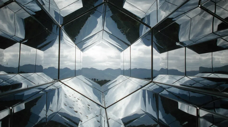Geometric mirrored panels reflect a cloudy sky and mountains, creating a kaleidoscopic visual effect.