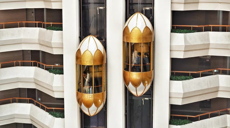 Two gold and white glass elevators ascend inside a multi-level modern atrium with terraces and plants.