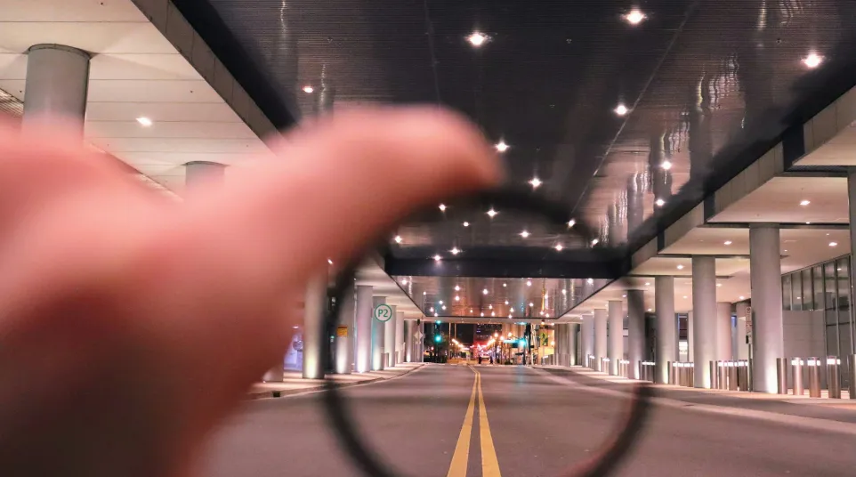 Hand holding a lens focusing on a city street under a modern, lit ceiling with pillars at night.
