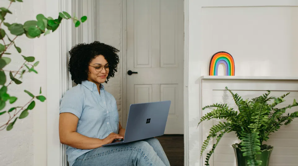 Woman with curly hair and glasses sitting on floor working on laptop in bright room with rainbow decor and green plant.