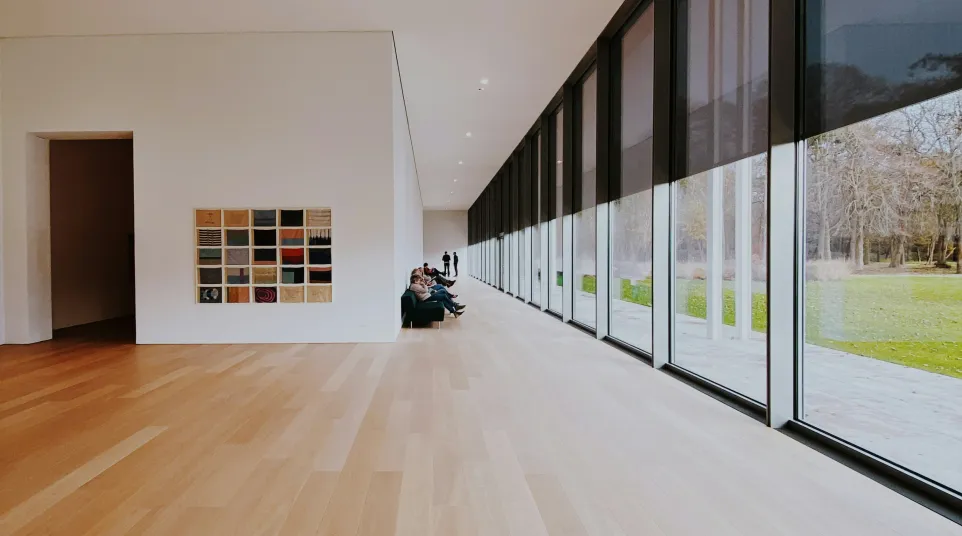 Minimalist gallery space with wooden floors, large windows with blinds, and people seated on a bench.
