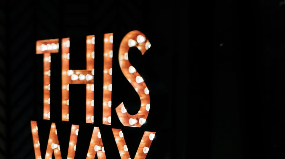 Illuminated marquee sign with words THIS WAY and arrow, reflecting on shiny black floor in dark room.