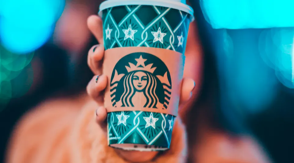 Hand holding a green Starbucks holiday cup with a blurred colorful bokeh background and person in orange coat.