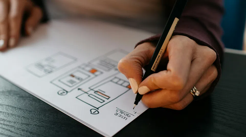 Close-up of a hand sketching a website user flow chart with a pen on white paper at a desk.