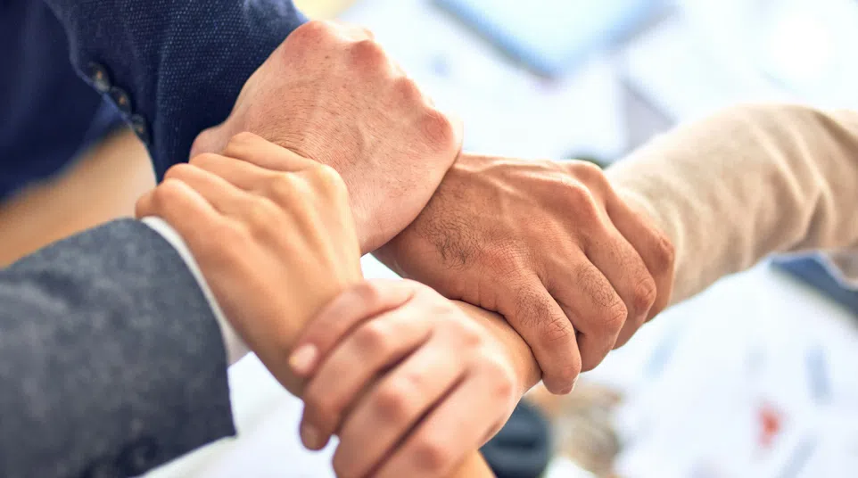 Four diverse business professionals joining hands in a teamwork gesture over an office desk with documents.
