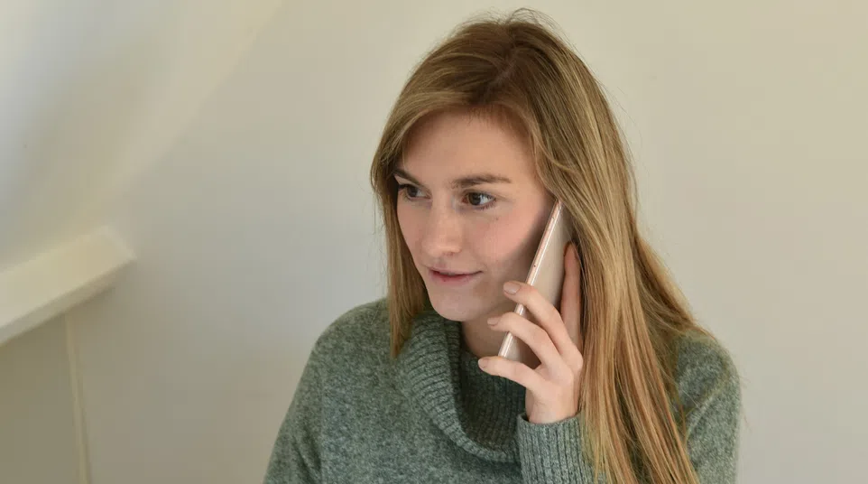 Woman in green sweater talking on phone while working at desk with computer in bright office space