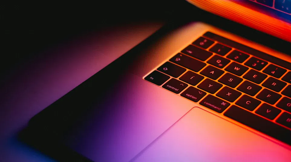 Partial view of a laptop keyboard illuminated by colorful neon lights in a dark setting.