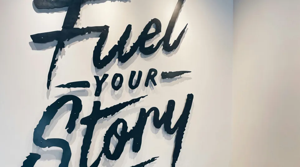 Cozy corner with two black leather chairs and wooden table under a wall sign saying Fuel Your Story in bold lettering.