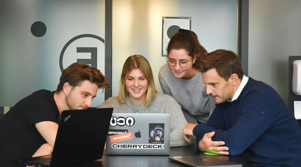 Four young professionals gathered around laptops collaborating in a modern office setting.
