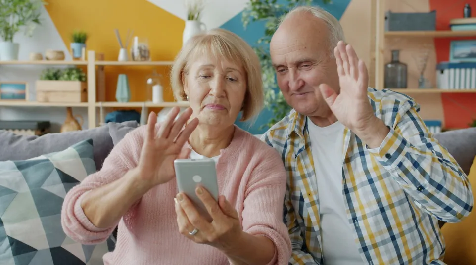 Senior couple video calling on smartphone waving hands in colorful living room.