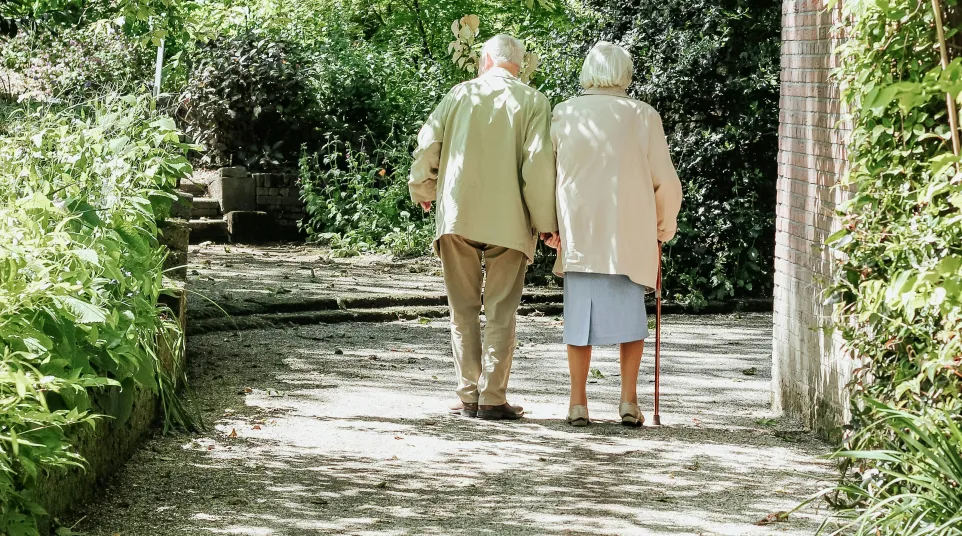 Elderly couple walking hand-in-hand on a shaded garden path surrounded by greenery.