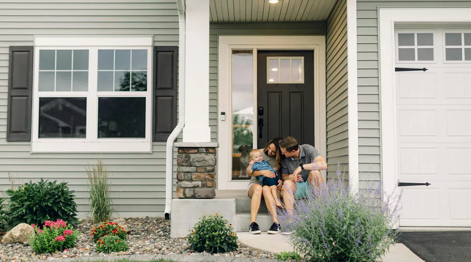 Family with toddler sitting on front porch steps of modern suburban house with stone accents and garden.