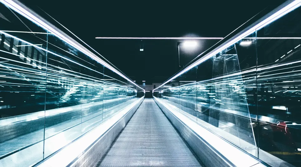 Futuristic illuminated walkway with glass railings and bright linear lights in a modern urban setting at night.