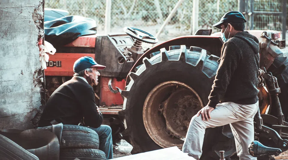 Two men wearing caps work beside a large red tractor in a rustic garage with old tires and tools.