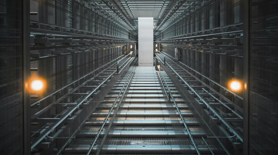 View looking up inside a modern glass elevator shaft with metal framework and warm lights illuminating the space