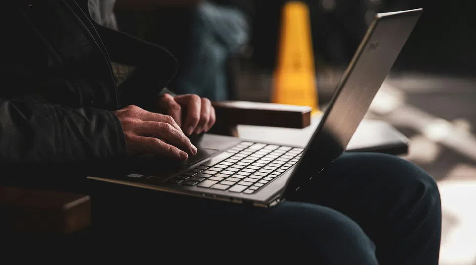 Person typing on a laptop keyboard while sitting in a dimly lit area with a blurred background.