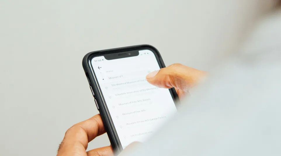 Person holding smartphone with location search results visible on screen in a minimalistic setting.