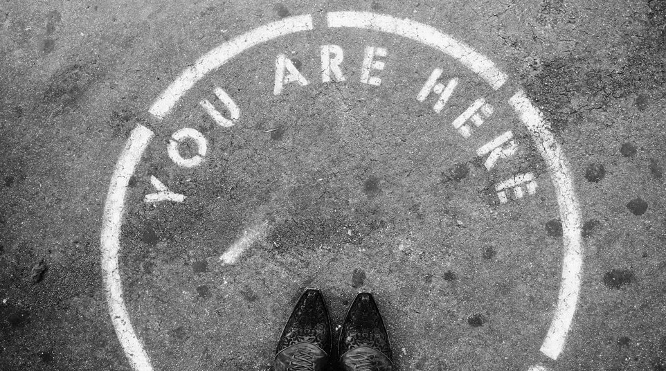 Black and white photo showing feet inside a painted circle on pavement with words You Are Here.