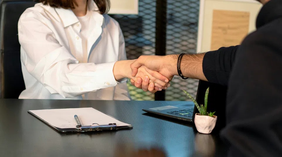 Two people shaking hands across a desk with a clipboard and laptop, symbolizing agreement or interview.