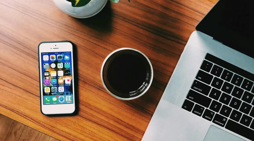Top view of a wooden desk with a smartphone, coffee cup, laptop, and a green potted plant with someone wearing jeans and sneakers.