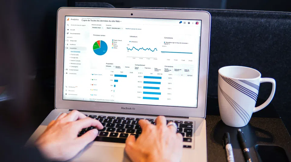 Person using a MacBook Air showing web analytics dashboard with charts, next to a striped coffee mug and pens.