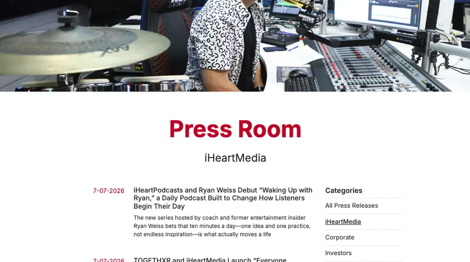 Radio host Enrique Santos speaking into microphone in iHeart Latino studio with audio equipment and drum set visible