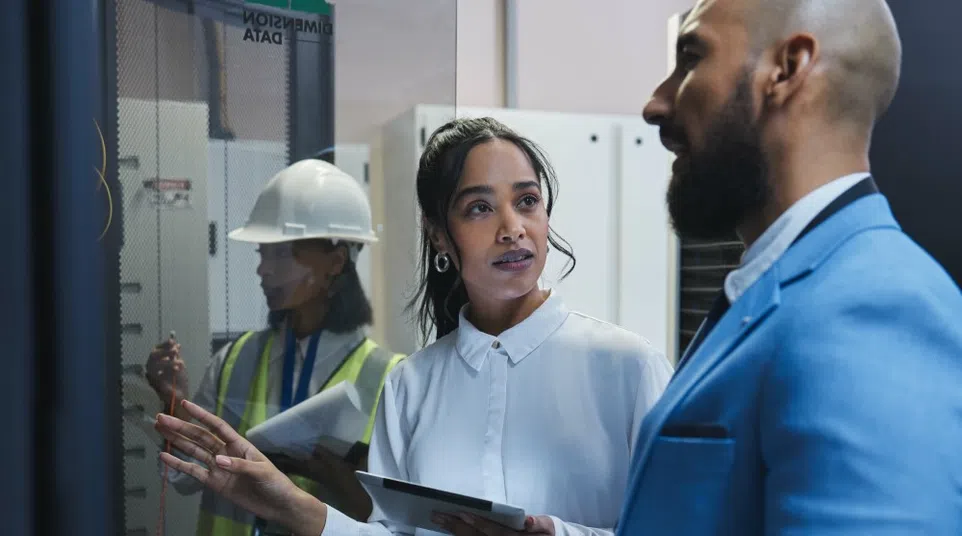 Two professionals discussing data on a screen with a worker in safety gear in the background inside a control room.
