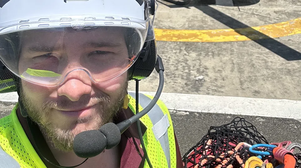 A worker wearing safety gear and headset stands near a red and white helicopter on a helipad under blue sky.
