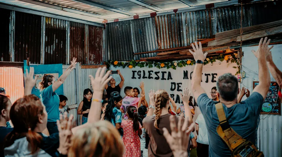 People raising hands in a lively church gathering inside a rustic metal building with a sign reading Iglesia Monte de Gracia.