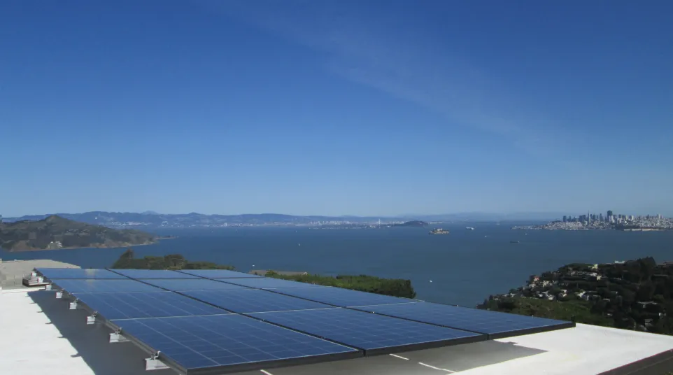 Solar panels installed on a sausalito rooftop overlooking a bay with a city skyline in the background under a clear blue sky.