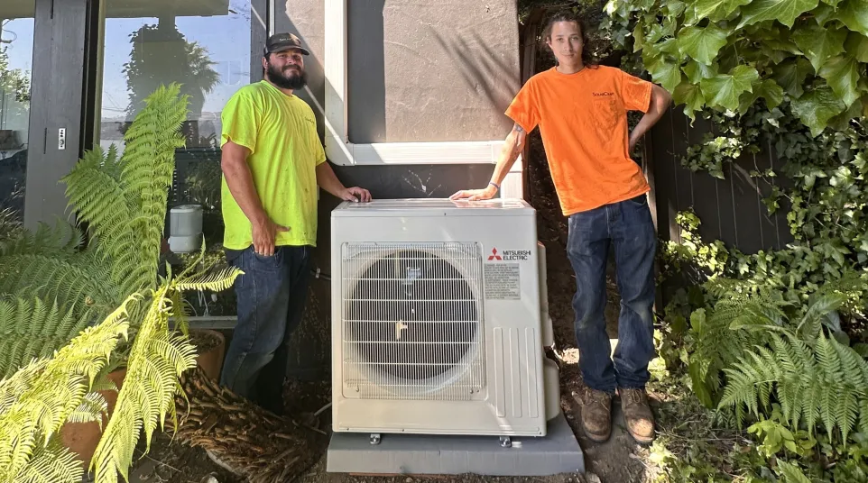 Two workers standing beside a newly installed Mitsubishi Electric outdoor HVAC unit outside a house surrounded by plants.