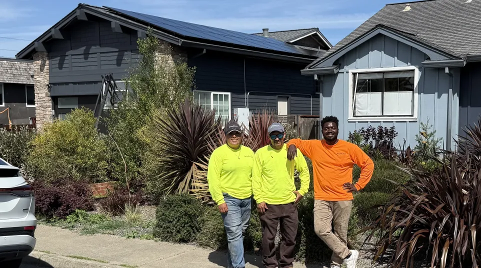 Three workers in bright safety shirts standing on a sidewalk by houses with garden plants and solar panels on the roof in larkspur