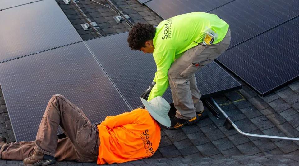 Two workers installing solar panels on a roof with safety gear and bright work shirts in daylight.