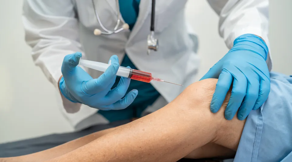 Doctor wearing gloves injecting a syringe into a patient's bent knee for treatment or pain relief.
